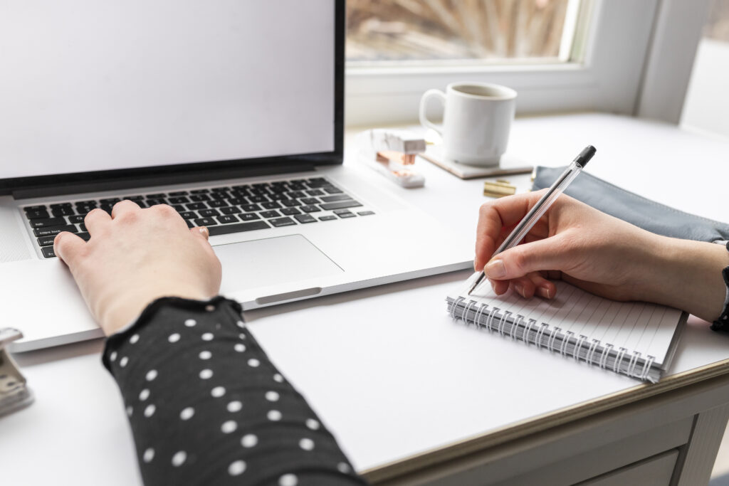 Person writing on notebook while using a laptop on an office setting.